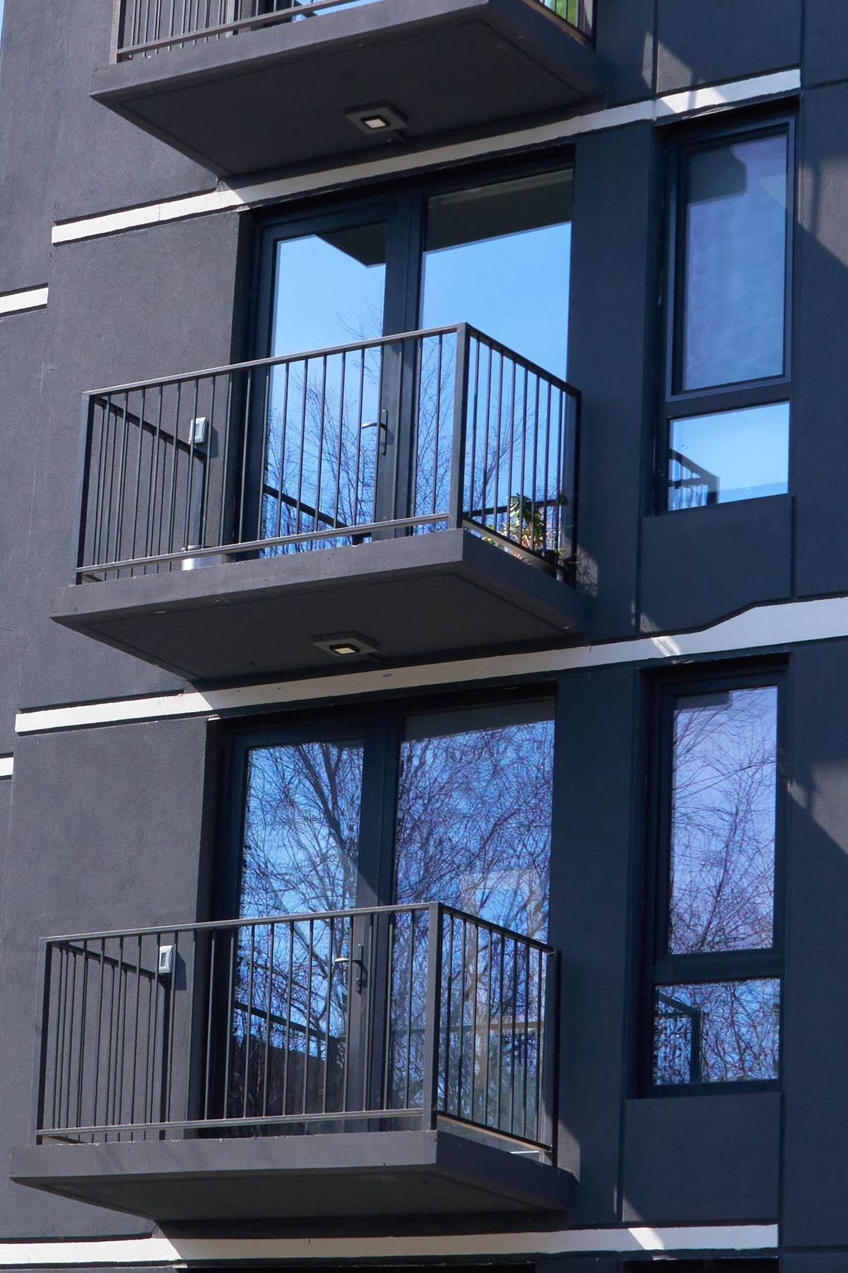 Two stacked balconies on a dark facade, each with a black terrace door and side light reflecting the sky
