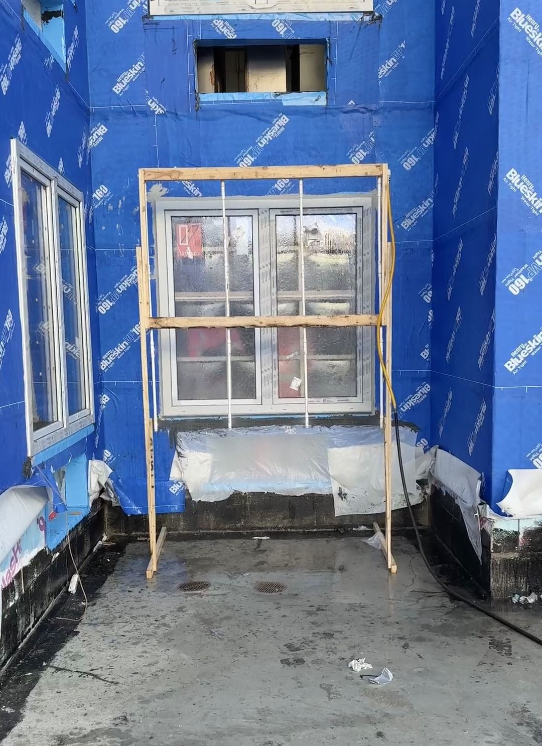 A spray rack on a wooden stand in front of an installed window in a wall still wrapped in its blue weather barrier, with water running down the glass