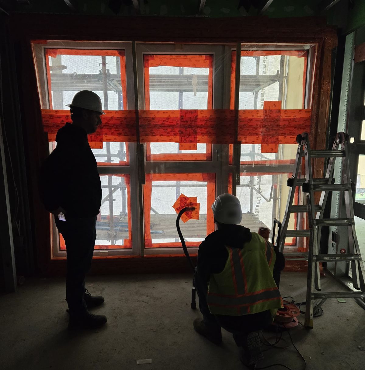 A field water test seen from inside a building under construction: a window taped up for the test, one person standing watching it and a technician kneeling at the hose