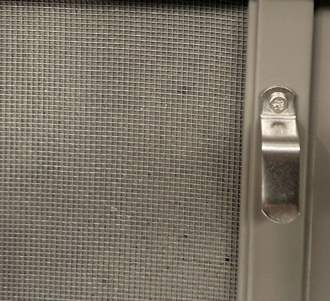 Close-up of an insect screen: fine charcoal mesh in a gray aluminum frame, with a spring-loaded metal fastener screwed to the side of the frame