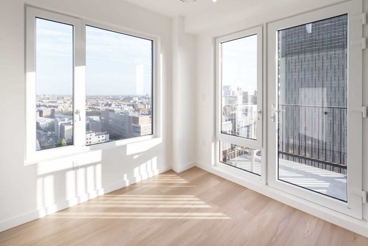 A sunlit empty corner room with a full-height terrace door and guardrail beside a three-light window
