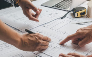 Blueprints on a table with people’s hands pointing and holding a pen—courtesy of Vistaza.