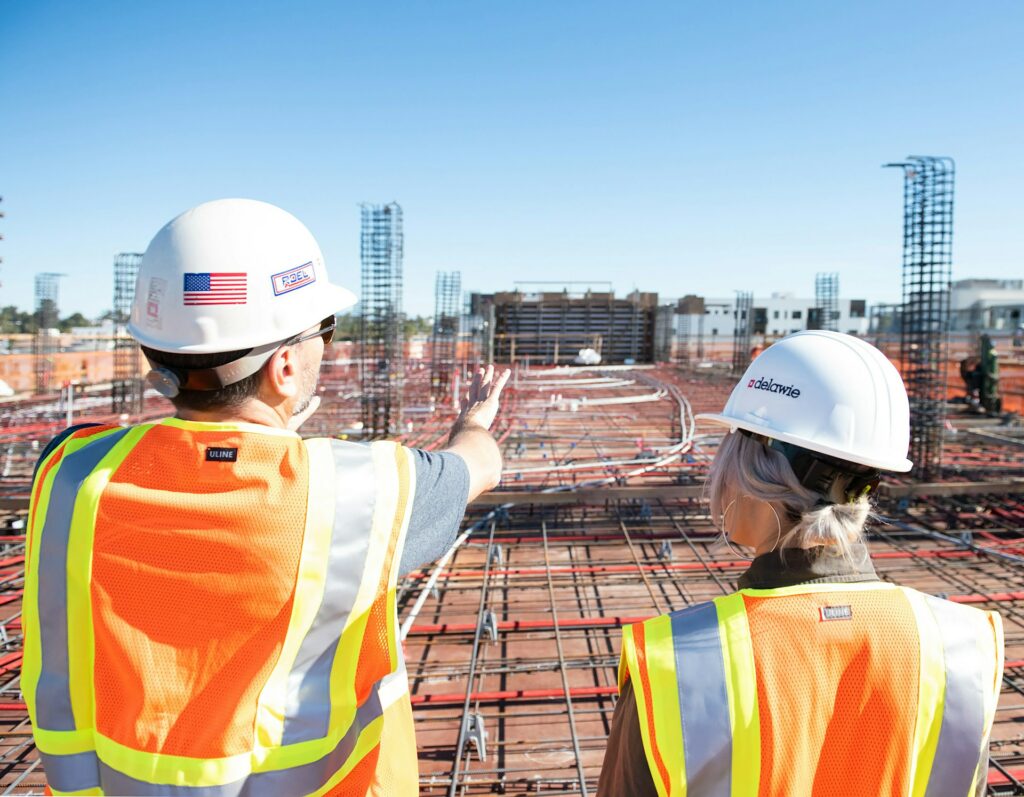Outdoor construction scene with two men holding blueprints and workers, rebar rods, and construction equipment in the background — photo by Vistaza.