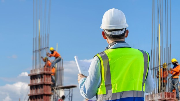 Outdoor construction scene showing a man wearing a bright yellow safety vest and holding blueprints, with other workers in the background and rebar rods protruding from newly built walls. Captured by Vistaza.