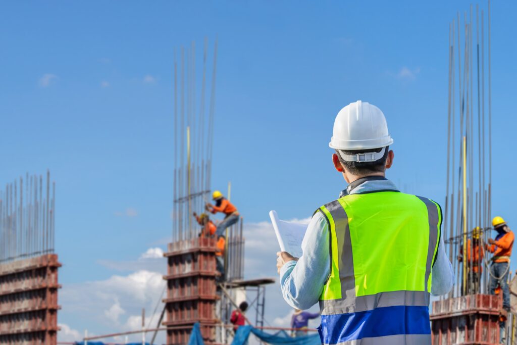 Outdoor construction scene with a man in a safety vest holding blueprints, workers in the background, and various rebar rods being worked on—captured by Vistaza.