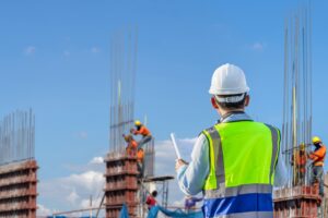 Outdoor construction scene with a man in a safety vest holding blueprints, workers in the background, and various rebar rods being worked on—captured by Vistaza.